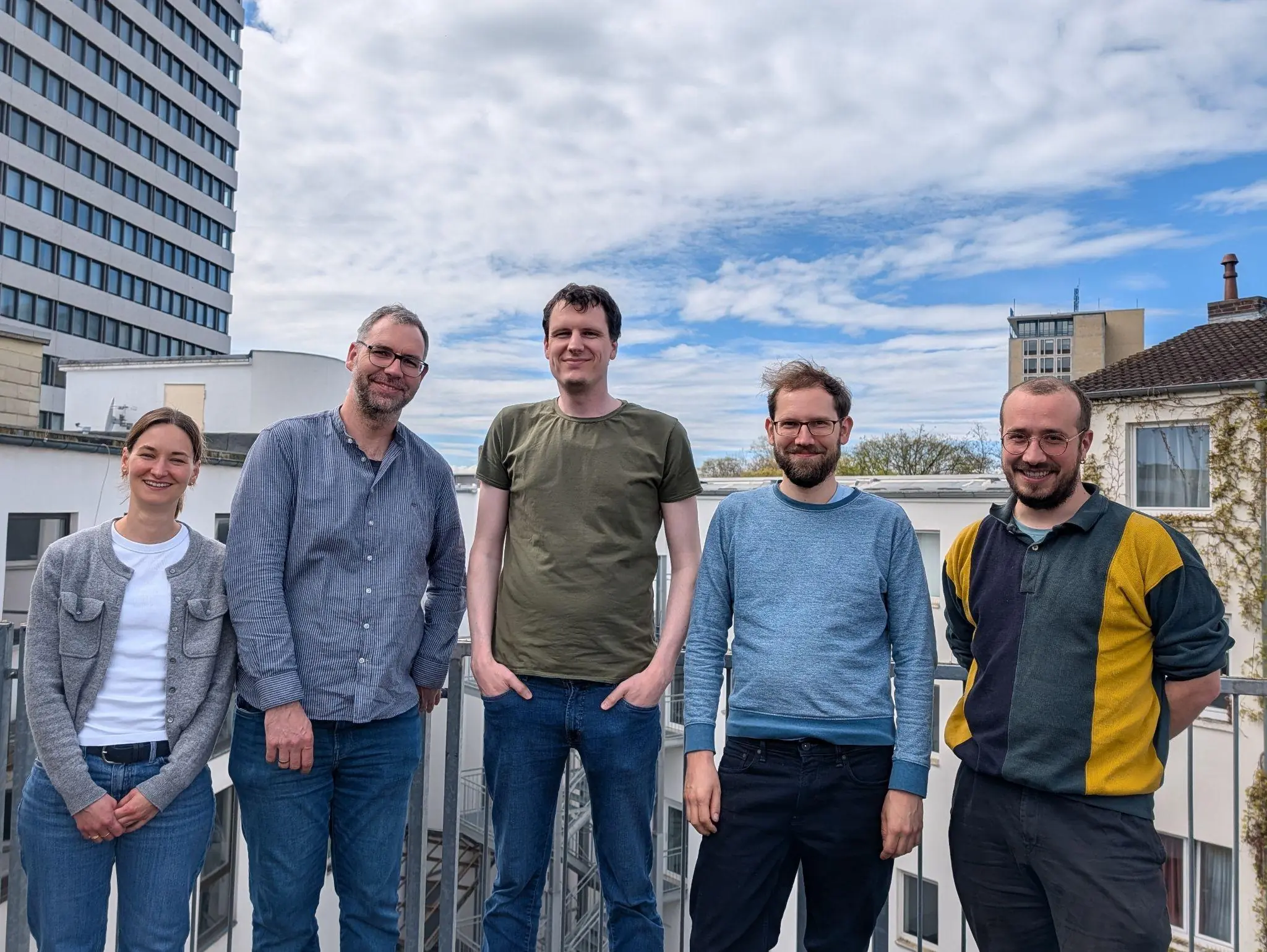 Five people smiling, standing shoulder to shoulder on a rooftop. Behind them is blue sky with clouds.