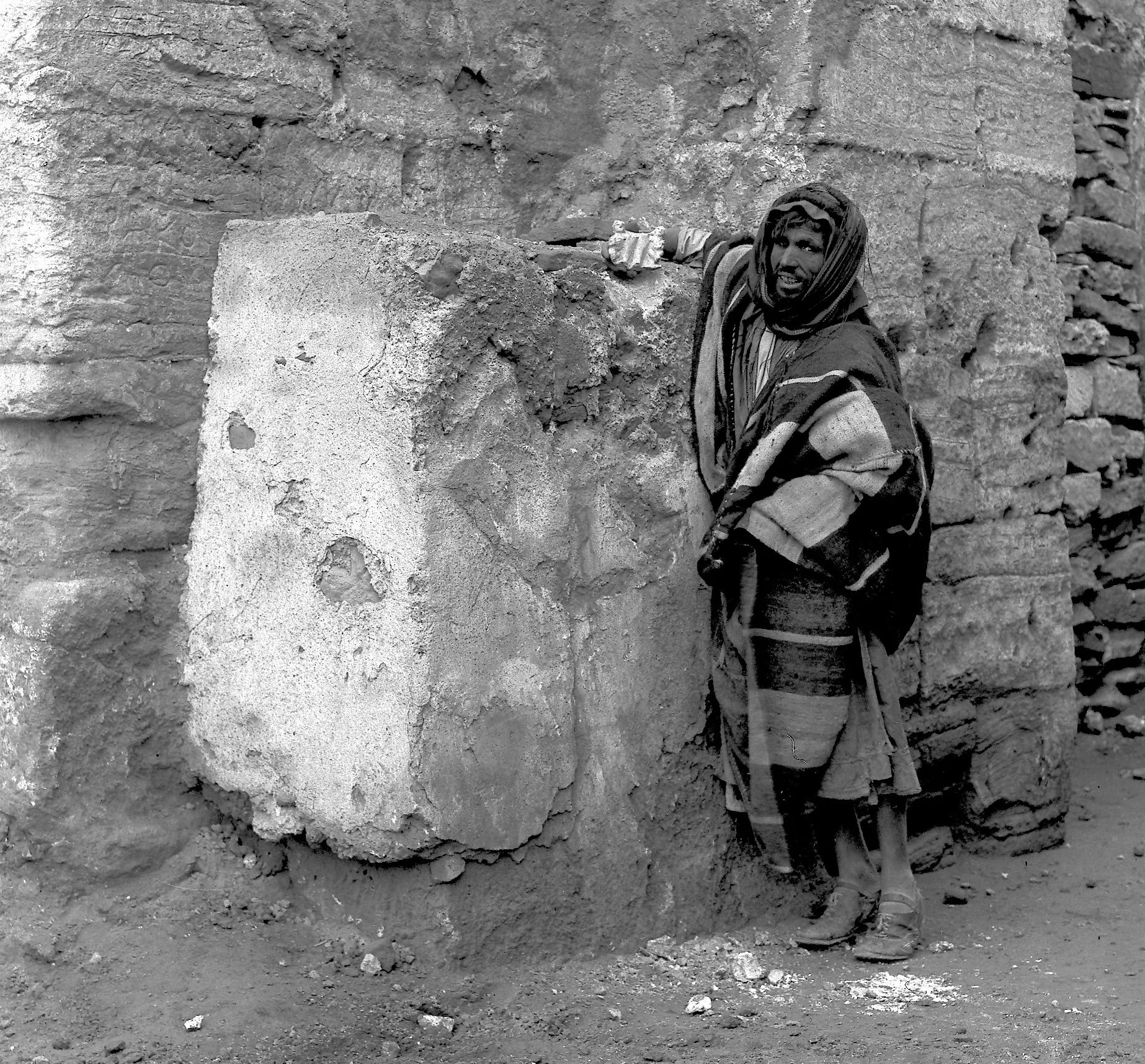 A black-and-white photo of a man wearing desert-appropriate clothing standing next to a stone altar carved into a wall of stone