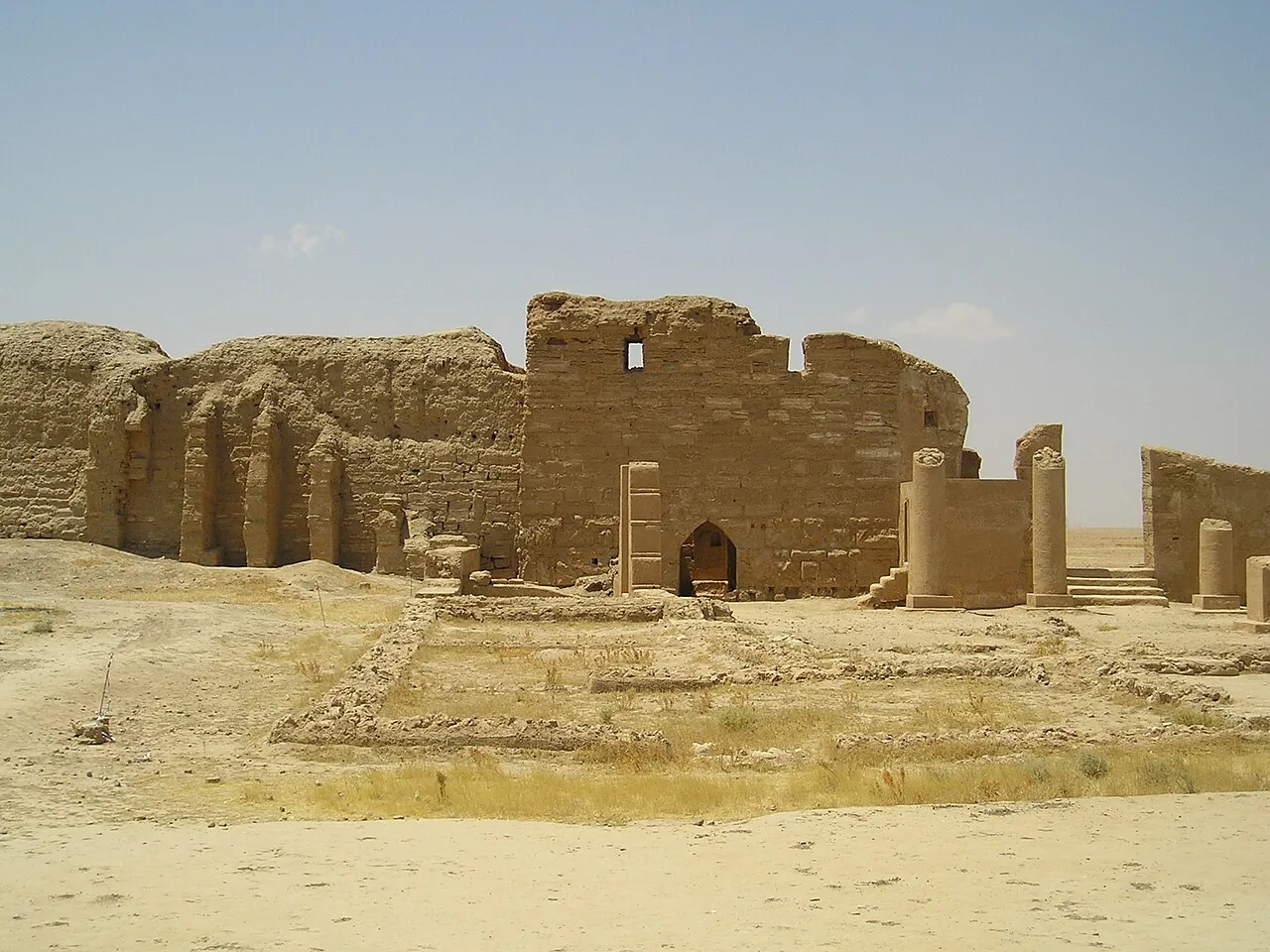 temple ruins with columns, stairs and a small entrance, framed by blue sky above and desert sands and grass below.