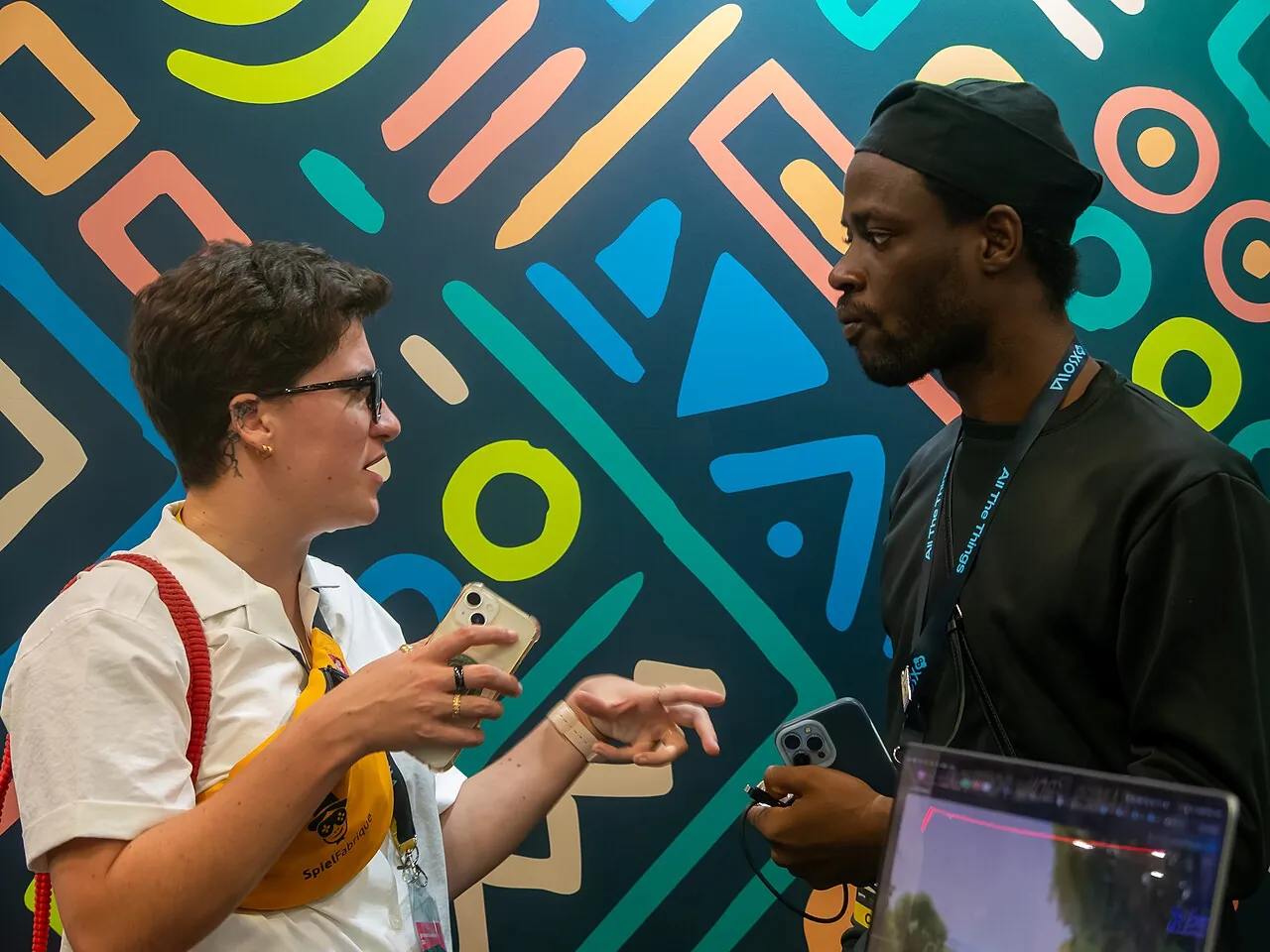 Two people in profile engage in a discussion in front of the brightly colored backdrop of the "Games from Nigeria" stand at gamescom 2026.